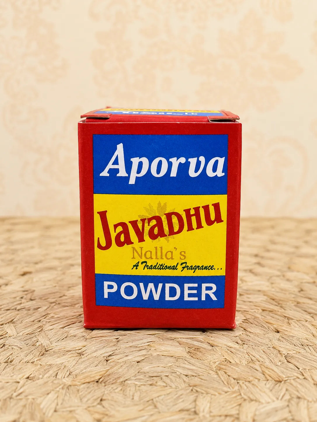 A red rectangular box of Javadhu Aporva powder fragrance, featuring a yellow and blue label, set on a woven straw mat against a patterned beige background.
