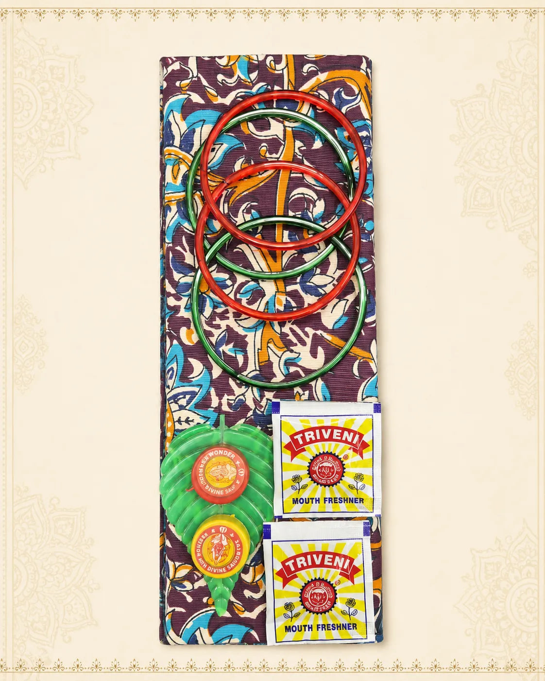 A colorful traditional Indian toy set featuring a patterned fabric board, red and green rings, a green and red ball, and two Triveni mouth freshener packets, arranged on a cream-colored patterned background.