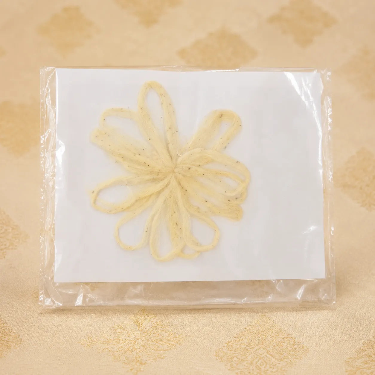A pale yellow, looped decorative hair accessory or comb piece displayed on a white backing card inside a clear plastic sleeve, set against a beige patterned fabric background.