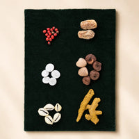 A rectangular black felt or fabric panel laid flat on a light beige background, decorated with small natural objects arranged in a grid-like pattern: red berries, two tan dried dates or nuts, white and brown stones, two white cowrie shells, and a piece of yellow ginger.