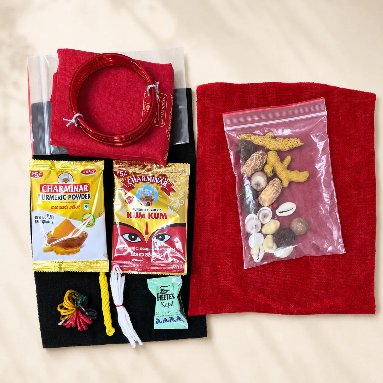 A flat-lay view of a traditional Indian matchmaking or ritual kit on a beige fabric background. The kit includes a red felt cloth, a red coiled cord, a black felt cloth with colorful tassels, two packets of Charminar Turmeric Powder, a packet of KJM Kum, a small packet of Hetta Clay, and a clear plastic bag containing various nuts, seeds, and spices.