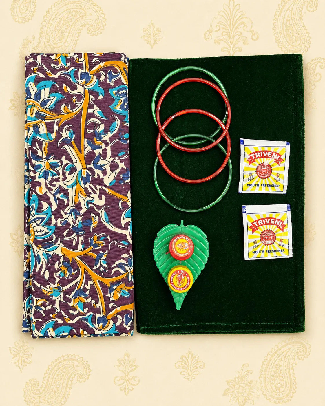 A set of traditional Indian items displayed on a beige patterned background: a folded multicolor patterned cloth on the left and a dark green velvet pouch on the right holding two red and green rings, a green leaf-shaped pendant with red and yellow circular details, and two small Triveni brand packets.